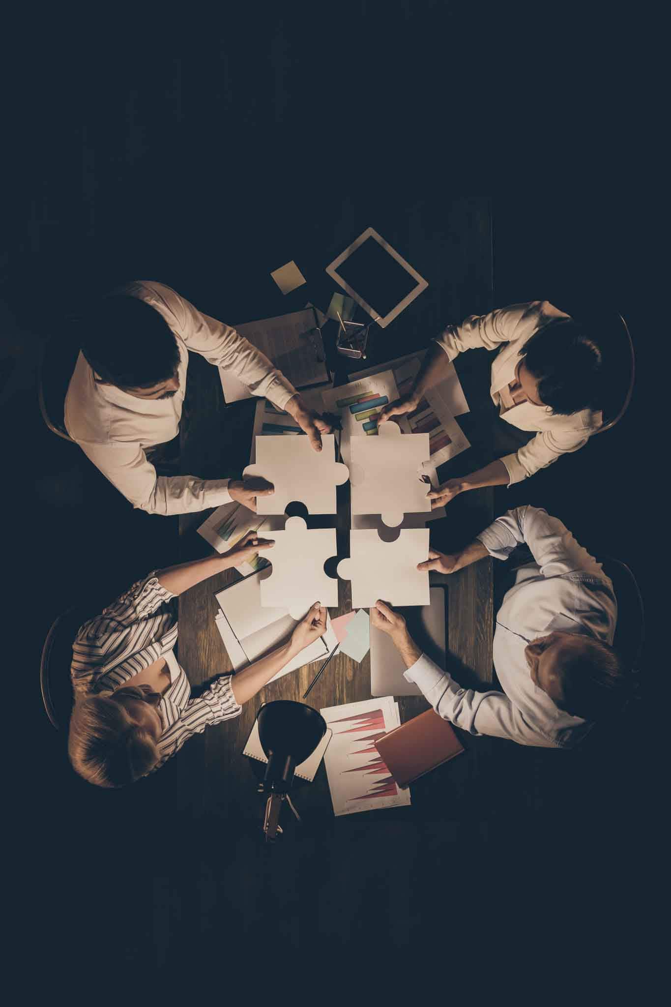 High angle above view vertical photo of four business people colleagues, sitting table circle working late night holding paper puzzle pieces find unity formalwear indoors